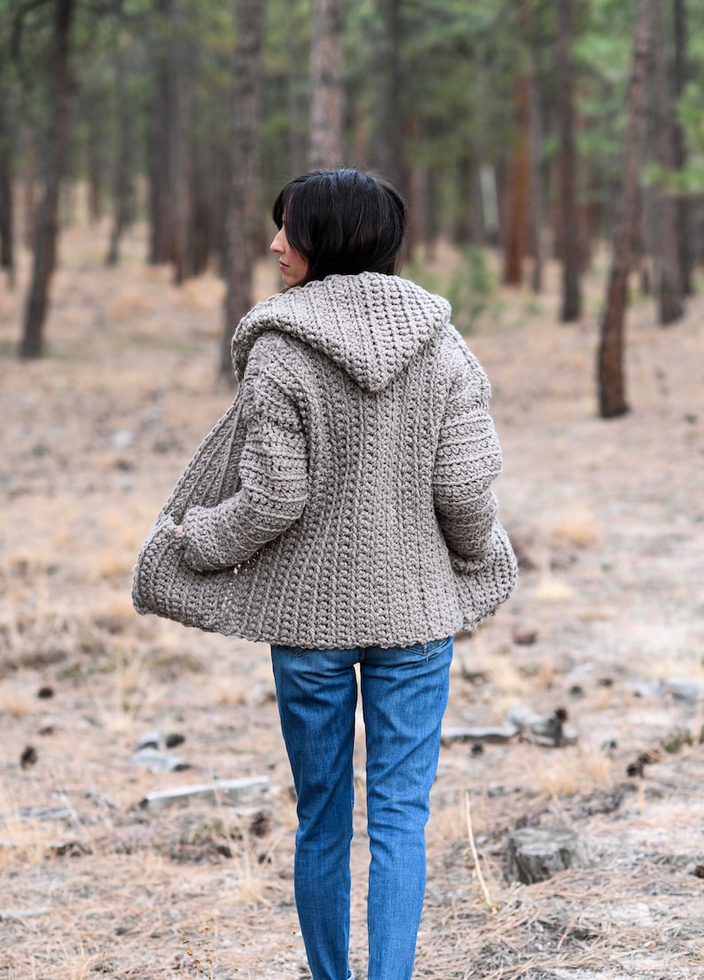 May include: A hooded cardigan in a light brown color, worn by a person in a forest setting. The cardigan has a relaxed fit and a long, flowing style.
