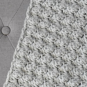 May include: A close-up of a grey crocheted blanket with a textured pattern.