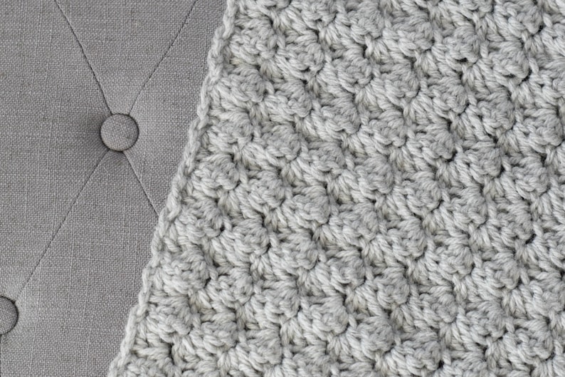 May include: A close-up of a grey crocheted blanket with a textured pattern.