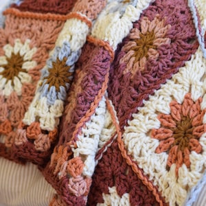May include: A crocheted throw blanket with a granny square pattern in shades of brown, cream, and pink. The blanket features a floral design with a central brown circle surrounded by cream and pink petals.