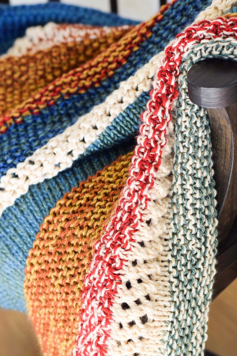Pu&ograve; includere: Una coperta di maglia a righe con un bordo rosso, bianco e blu. La coperta &egrave; realizzata con filati di diversi colori, tra cui blu, verde, marrone e bianco.