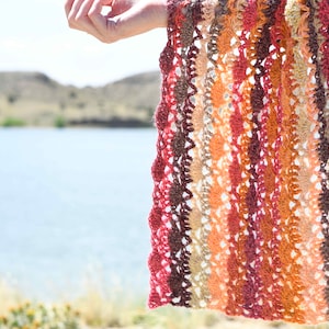 以下が含まれることがあります： 茶色、オレンジ、赤の色合いのストライプ模様のクロシェ編みショール。繊細な透かし編みのステッチでできています。