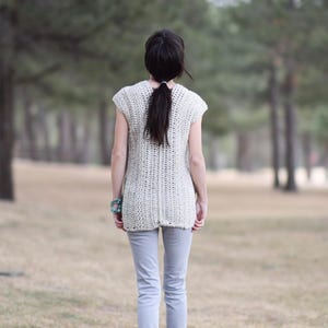 Puede incluir: Una mujer con un top de crochet blanco de manga corta y pantalones vaqueros azules claros. Est&aacute; de pie en un campo de hierba con &aacute;rboles en el fondo.