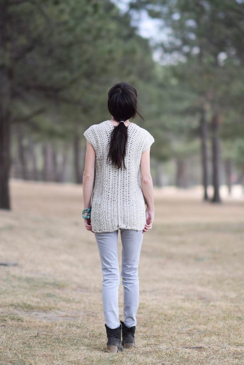 Puede incluir: Una mujer con un top de crochet blanco de manga corta y pantalones vaqueros azules claros. Est&aacute; de pie en un campo de hierba con &aacute;rboles en el fondo.