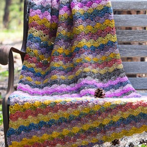 May include: A crocheted blanket featuring a wavy, multi-colored pattern in shades of mustard yellow, magenta, blue, and grey. The yarn has a textured, speckled appearance throughout the design. A single small brown pinecone rests on the blanket.