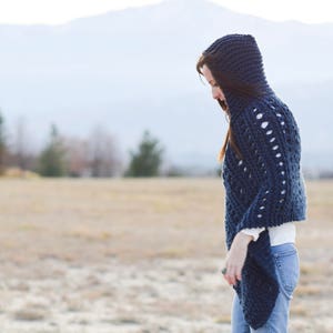 May include: A navy blue hooded knitted poncho with a lacy pattern. The poncho is worn by a person standing in a field.