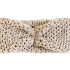 Puede incluir: Diadema beige tejida a mano con un diseño de nudo retorcido. La diadema está hecha de un material de punto grueso y es de color neutro. El artículo se muestra sobre un fondo blanco.
