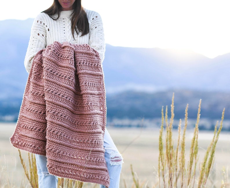 May include: A woman is holding a crocheted blanket in a dusty rose color. The blanket has a textured stitch pattern and is draped over her arm. She is standing in a field with tall grass and mountains in the background.