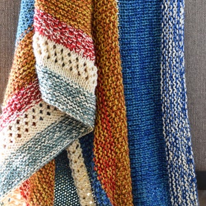 May include: A multi-coloured knitted throw blanket with stripes of blue, beige, red, and orange. The blanket is folded and draped over a sofa.