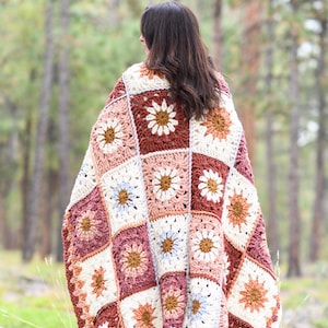 May include: A crocheted blanket with a floral pattern in shades of brown, pink, white, and blue. The blanket is made of squares with a daisy design in the center of each square. The blanket is draped over a person standing in a forest.