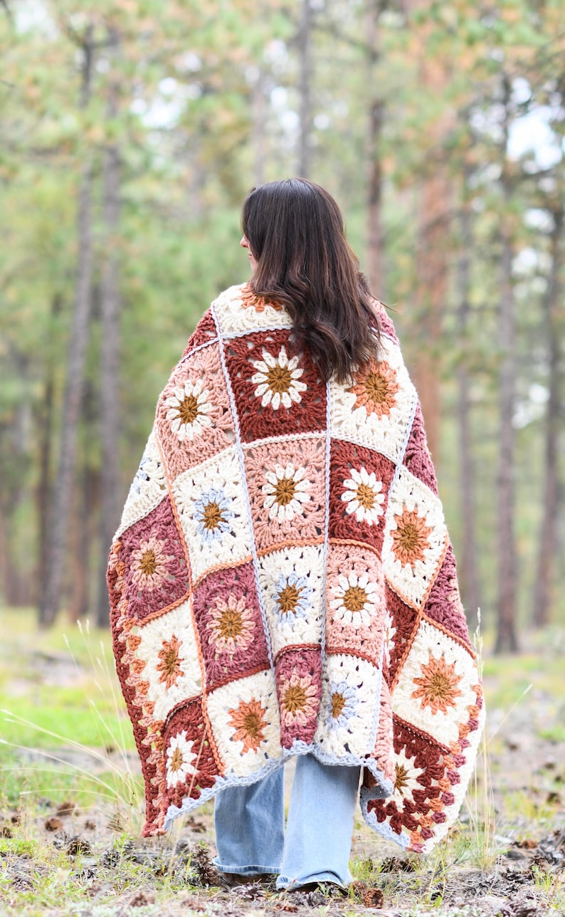 May include: A crocheted blanket with a floral pattern in shades of brown, pink, white, and blue. The blanket is made of squares with a daisy design in the center of each square. The blanket is draped over a person standing in a forest.