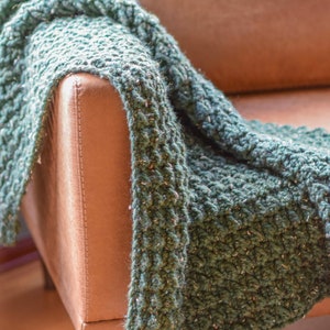 Op de afbeelding: Een groene gebreide deken gedrapeerd over een bruine leren fauteuil. De deken heeft een textuur, grof gebreid patroon.