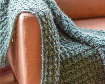 May include: A green knitted throw blanket draped over a brown leather armchair. The blanket has a textured, chunky knit pattern.