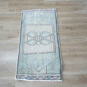 May include: A rectangular area rug featuring a faded mint green and cream color palette. The rug displays a central geometric floral motif enclosed within a square border, with additional linear patterns and small diamond shapes at the top and bottom.