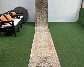 May include: A long, narrow, beige runner rug with a geometric pattern in shades of brown, pink, and white. The rug has a fringed edge and is hanging on a wall.