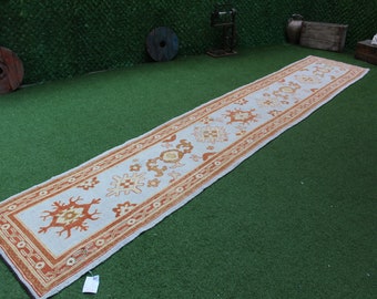 May include: A long, white runner rug with an orange and cream geometric pattern. The rug is laid out on a green artificial grass surface.