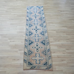 May include: A long, narrow runner rug featuring a light beige base color with a repeating pattern of blue geometric cross shapes and diamond motifs. The rug has a dark blue border with intricate triangular and diamond-shaped designs.