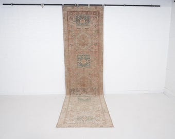 May include: A vintage runner rug with a faded, distressed appearance. The rug features a central design with geometric patterns in shades of brown, beige, and teal. The rug is displayed against a white wall and floor.