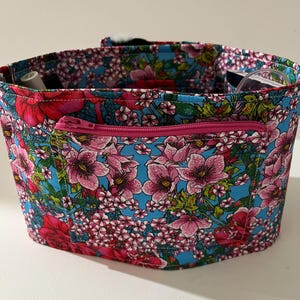 Könnte beinhalten: Eine Tragetasche mit floralem Muster und einer rosa Reißverschlusstasche. Die Tasche hat ein lebendiges Muster aus rosa Blumen und weißen Blüten auf türkisfarbenem Hintergrund. Das Innere der Tasche ist sichtbar und scheint kleine Gegenstände zu enthalten.