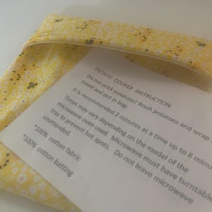 Puede incluir: Una bolsa de tela amarilla para cocinar patatas con un patrón de flores y abejas blancas. Encima hay una hoja de instrucciones blanca que dice: POTATO COOKER INSTRUCTION: Do not prick potatoes! Wash potatoes and wrap in towel and put in bag. 100% cotton fabric, 100% cotton batting.