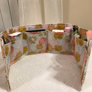 Puede incluir: Un organizador de tela rosa y amarillo con múltiples compartimentos. La tela tiene un estampado de piña. El organizador está vacío, excepto por algunos artículos en los compartimentos.
