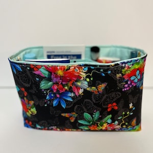 Könnte beinhalten: Ein schwarzer Stofforganizer mit einem Blumen- und Schmetterlingsmuster. Der Stoff ist mit einem hellblauen Stoff gefüttert. Der Organizer ist mit verschiedenen Gegenständen gefüllt, darunter eine blau-weiße Schachtel, eine weiße Schachtel und eine schwarze Bürste.