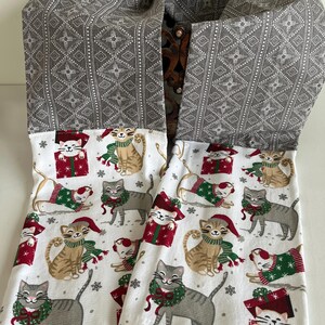 以下が含まれることがあります： 白地のキッチンタオル2枚。お祝いの猫のプリントが施されています。猫はサンタ帽、スカーフ、リースを身につけ、雪の結晶とギフトボックスに囲まれています。タオルはグレーの幾何学模様でトリミングされています。