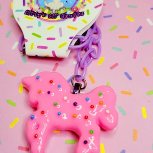 May include: A pink, glossy unicorn-shaped charm with colorful sprinkles, attached to a purple chain. The charm is connected to a tag with a cat illustration and the text "Nifty's Lil Krafts" on a background of colorful sprinkles.