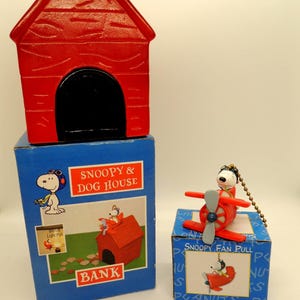 May include: A red Snoopy dog house bank with a black door sits atop a blue box. A Snoopy fan pull in a red airplane is on a blue box. The boxes feature Snoopy graphics and the text "Snoopy & Dog House" and "Snoopy Fan Pull".
