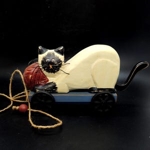 May include: A wooden toy cat with a cream-colored body, black and tan markings, and blue eyes. The cat is holding a red ball of yarn and is mounted on a blue cart with black wheels. A string with a red bead is attached.