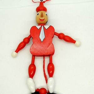 May include: A wooden marionette doll with a red outfit, including a hat and shirt with a white and blue tie design. The arms and legs are jointed, with white hands and feet, and black shoes. A red string is attached for control.