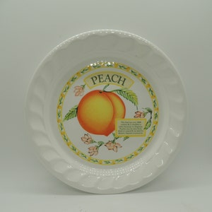 May include: White ceramic plate with a peach design. The plate has a scalloped edge and features a yellow and green border with pink flowers. The text "PEACH" is written in a yellow banner above the peach. The text below the peach reads: "This fruit has over 2000 varieties & is classified as a drupe. Peaches may be eaten fresh, dried or canned & are used for desserts,  squares or preserves."