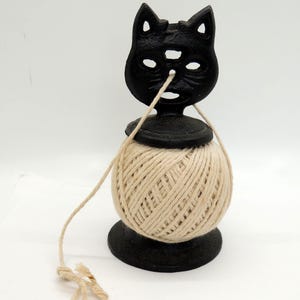 May include: A black cast iron twine holder featuring a stylized cat head design at the top. The holder supports a round ball of natural beige twine that feeds through the center of the cat's face. The base is circular and solid black.