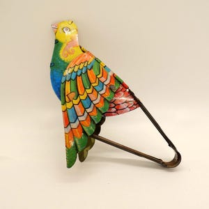 May include: A colorful metal bird toy with a yellow head, blue body, and vibrant wings in shades of orange, green, and red. The bird has a long, curved metal handle attached to its body, likely for display or play.