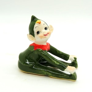 May include: A glossy ceramic figurine of an elf in a seated position, reaching forward to touch its feet. The elf has a dark green suit, a pointed green hat, a bright red collar, and a pale face with painted facial features and rosy cheeks.