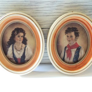 May include: Two oval framed portraits of children. The girl is wearing a blue and red dress with a white blouse and a necklace. The boy is wearing a blue vest with a white shirt and a red scarf. Both portraits have a light brown background.
