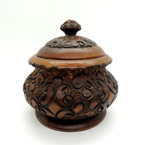 May include: A round, dark brown wooden lidded jar featuring intricate, deep-carved scrollwork patterns across the body and the lid. The container sits on a circular base and is topped with a small, carved knob handle on the center of the lid.