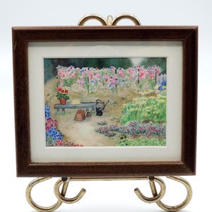 May include: A framed watercolor painting depicting a garden scene with a wooden bench, a small watering can, terracotta pots, and a trellis covered in pink and purple flowers. The artwork is enclosed in a dark brown wooden frame with a white mat.