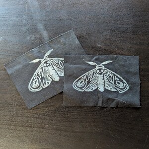May include: Two black fabric patches with a white moth design. The moth illustration features intricate details, including a skull-like pattern on the head and detailed wing patterns. The patches are rectangular and appear to be handmade.