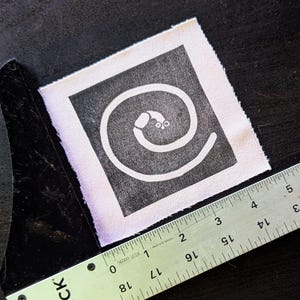 Puede incluir: Un parche de tela cuadrado con un dise&ntilde;o de espiral blanco sobre un fondo negro. El parche mide aproximadamente 5 cm de ancho. Una regla met&aacute;lica se coloca en diagonal sobre el parche, con el logotipo "BLICK" visible.