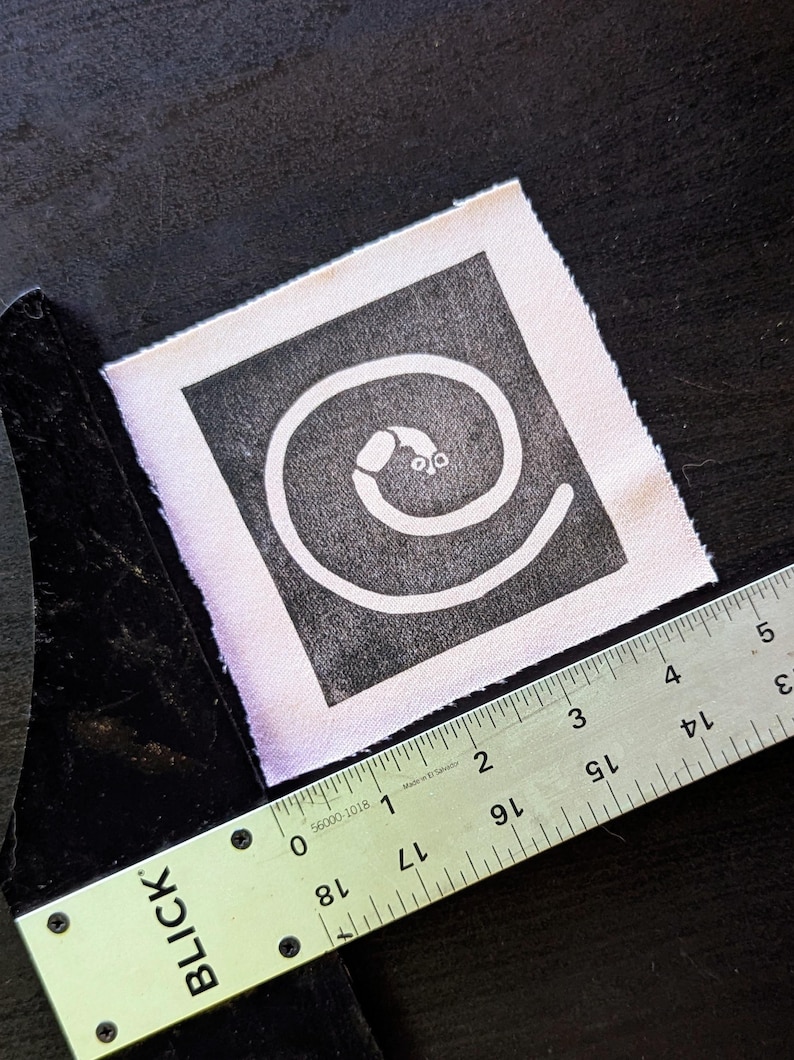 Puede incluir: Un parche de tela cuadrado con un dise&ntilde;o de espiral blanco sobre un fondo negro. El parche mide aproximadamente 5 cm de ancho. Una regla met&aacute;lica se coloca en diagonal sobre el parche, con el logotipo "BLICK" visible.