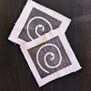 May include: Two square fabric patches with a black spiral design and a small detail at the end. The patches have a white border and are set against a dark background.