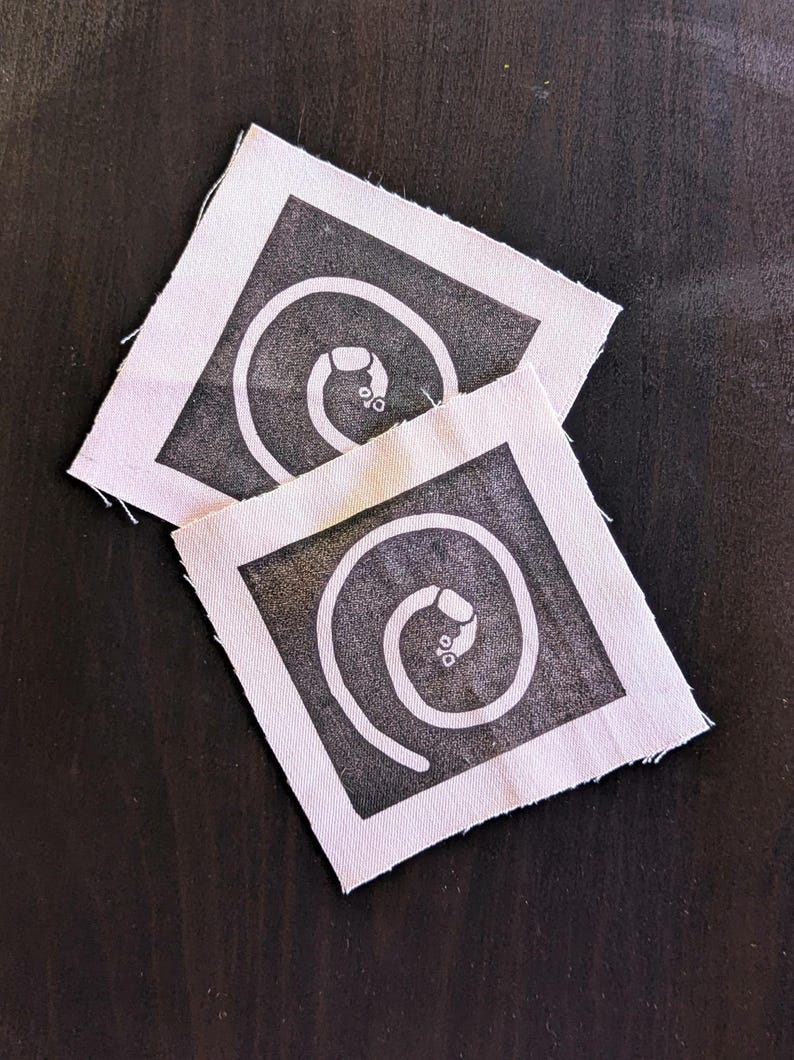 Puede incluir: Dos parches cuadrados de tela con un dise&ntilde;o de espiral negro y un peque&ntilde;o detalle en el extremo. Los parches tienen un borde blanco y est&aacute;n colocados sobre un fondo oscuro.