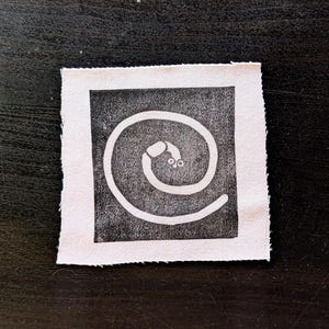 Puede incluir: Un peque&ntilde;o parche de tela cuadrado con un dise&ntilde;o en blanco y negro. El dise&ntilde;o es una espiral blanca con un peque&ntilde;o ojo estilizado en el centro, sobre un fondo cuadrado negro. El parche tiene un borde crudo sin terminar.