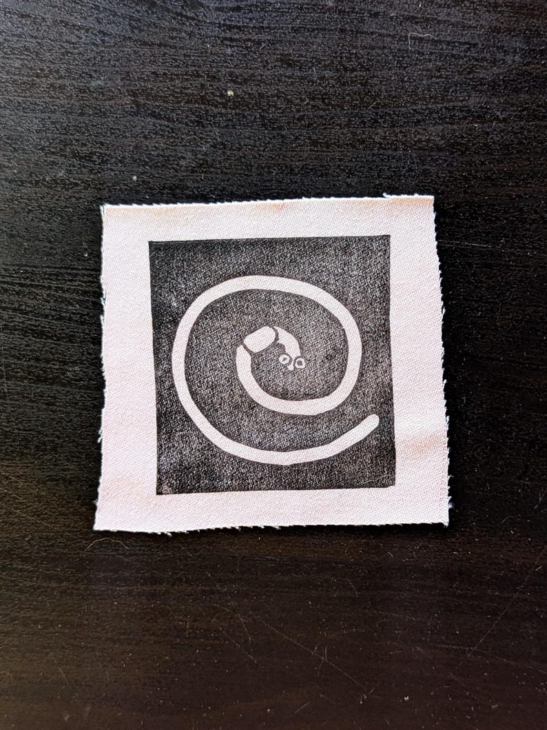 Puede incluir: Un peque&ntilde;o parche de tela cuadrado con un dise&ntilde;o en blanco y negro. El dise&ntilde;o es una espiral blanca con un peque&ntilde;o ojo estilizado en el centro, sobre un fondo cuadrado negro. El parche tiene un borde crudo sin terminar.
