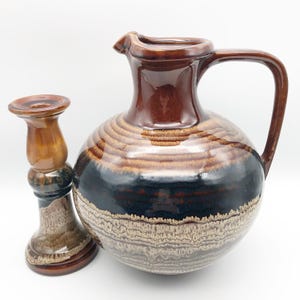 May include: A brown ceramic pitcher with a handle and a matching candlestick. The pitcher has a round body with a dark blue and tan pattern. The candlestick is a similar color and design.