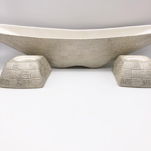 May include: A cream-colored ceramic centerpiece bowl with a textured, woven pattern. Two matching square candle holders are in front of the bowl. The bowl has a curved, boat-like shape, and the candle holders have a textured surface.