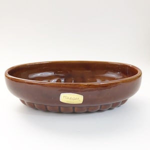 May include: A brown ceramic Haeger oval dish with a glossy finish. The dish has a scalloped edge and a small, rectangular sticker with the Haeger logo. The dish is empty and sits on a white surface.