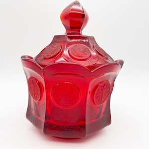 May include: A ruby red glass candy dish with a lid. The dish has a hexagonal shape with embossed circular designs. The lid is topped with a pointed finial. The glass has a glossy finish.
