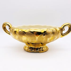 May include: A gold-toned ceramic bowl with a scalloped rim and two side handles. The exterior features a reflective metallic gold finish with a textured, mottled appearance, while the interior is a smooth, solid off-white color.